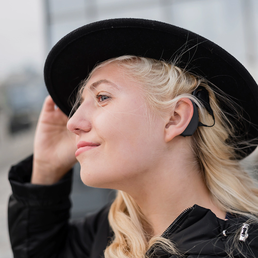 Uśmiechnięta kobieta w blond włosach z czarnym kapeluszem na głowie, ze zbliżeniem na pętle indukcyjną za uchem.
