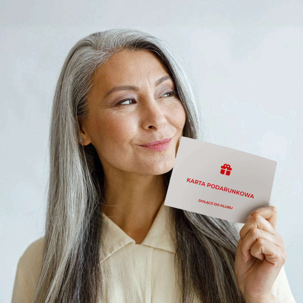 Starsza kobieta w siwych włosach patrząca w dal z uśmiechem na twarzy, w ręku trzyma białą kartę podarunkową z czerwonymi oznaczeniami \