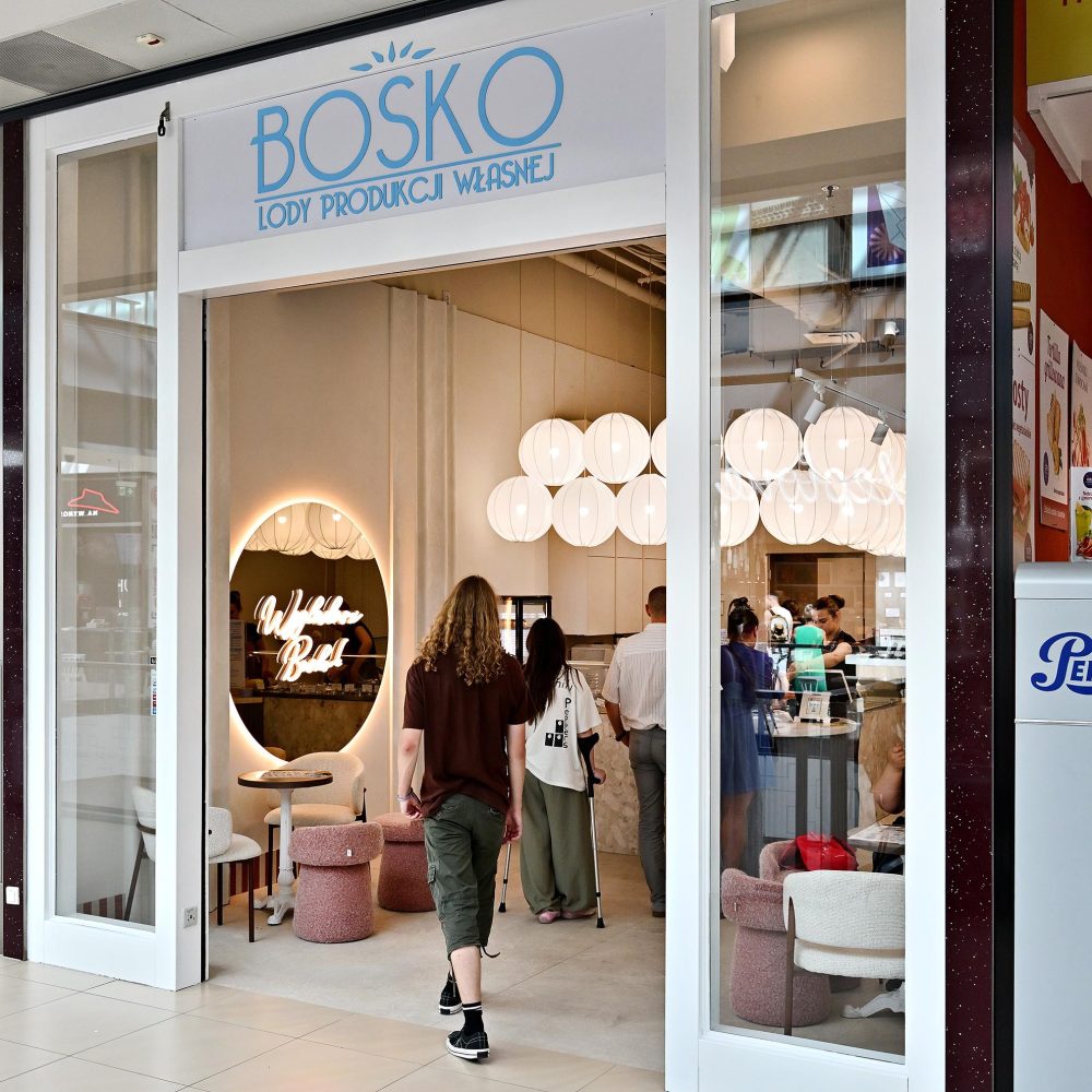 Wejście do lodziarni Bosko w galerii handlowej. W jasnym, eleganckim wnętrzu z dekoracyjnymi, okrągłymi lampami klienci stoją przy ladzie, a przed lokalem ustawiono różowe i kremowe siedziska oraz niewielkie stoliki. Na ścianie widoczne jest podświetlane lustro z neonowym napisem „Welcome Back”.