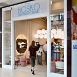 Wejście do lodziarni Bosko w galerii handlowej. W jasnym, eleganckim wnętrzu z dekoracyjnymi, okrągłymi lampami klienci stoją przy ladzie, a przed lokalem ustawiono różowe i kremowe siedziska oraz niewielkie stoliki. Na ścianie widoczne jest podświetlane lustro z neonowym napisem „Welcome Back”.