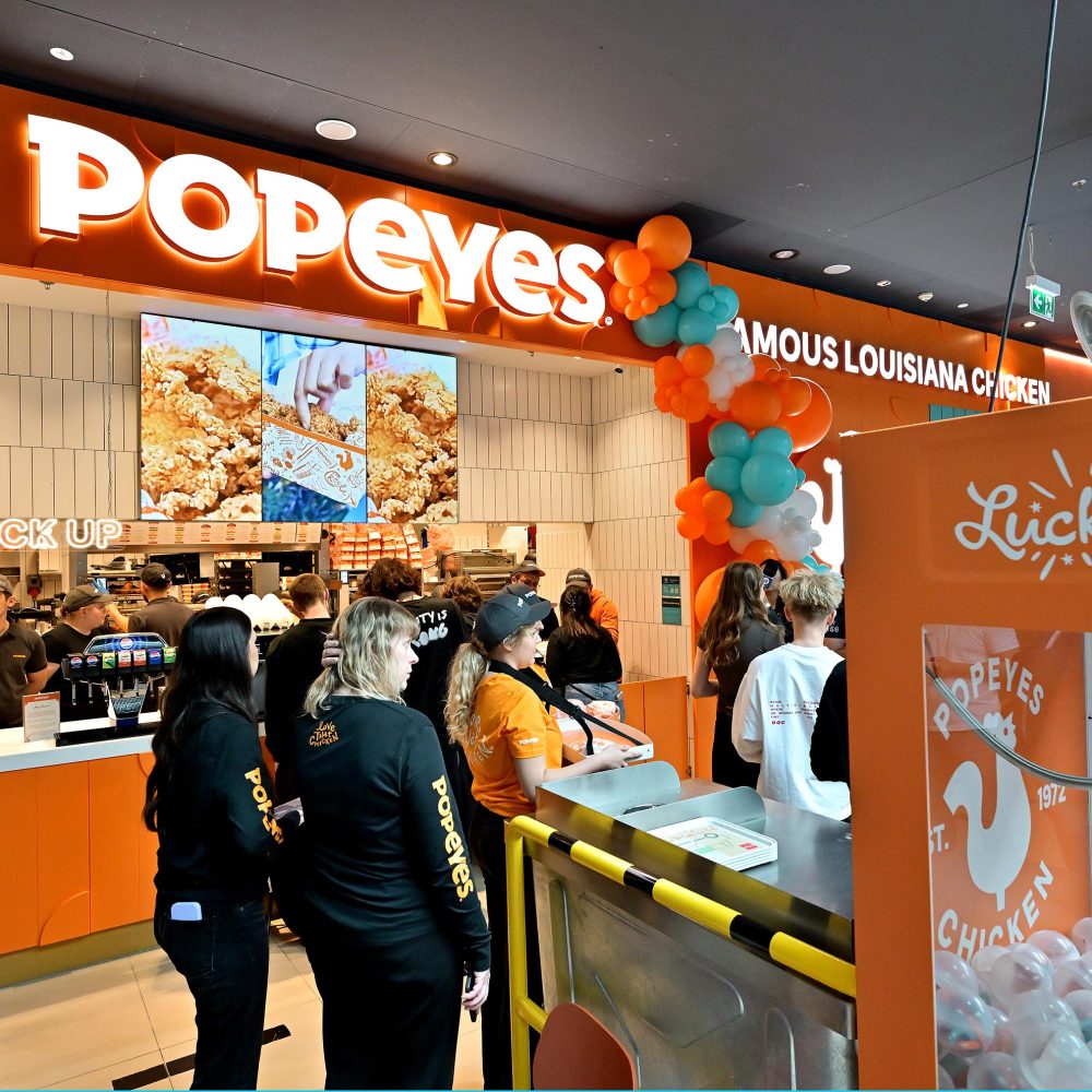 Wejście do restauracji Popeyes udekorowane pomarańczowo-turkusowym łukiem z balonów. Przed ladą stoi kolejka klientów, a pracownicy obsługują zamówienia.