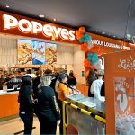 Wejście do restauracji Popeyes udekorowane pomarańczowo-turkusowym łukiem z balonów. Przed ladą stoi kolejka klientów, a pracownicy obsługują zamówienia.