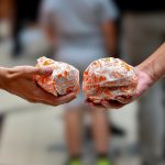 Zdjęcie przedstawia z bliska dwie pary rąk trzymające burgery z restauracji Popeyes zawinięte w charakterystyczny, biało-pomarańczowy papier z grafikami. Tło zdjęcia jest mocno rozmyte i widać na nim sylwetki przechodzących osób, co skupia całą uwagę na trzymanym jedzeniu.