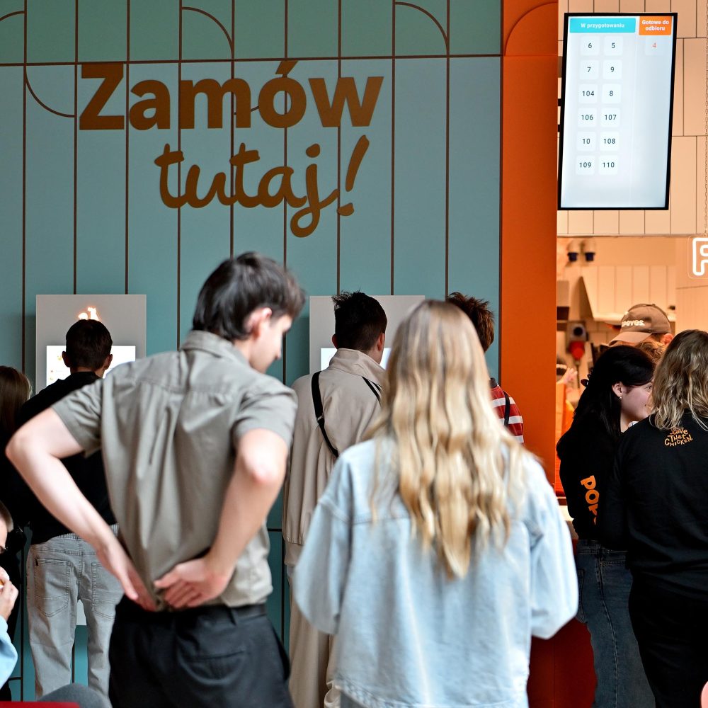 podświetlonym szyldem restauracji Popeyes. W tle widoczne są kolorowe dekoracje z balonów, pracownica w pomarańczowej koszulce oraz ekrany wyświetlające m.in. postać psa w barwnym stroju.