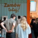 podświetlonym szyldem restauracji Popeyes. W tle widoczne są kolorowe dekoracje z balonów, pracownica w pomarańczowej koszulce oraz ekrany wyświetlające m.in. postać psa w barwnym stroju.