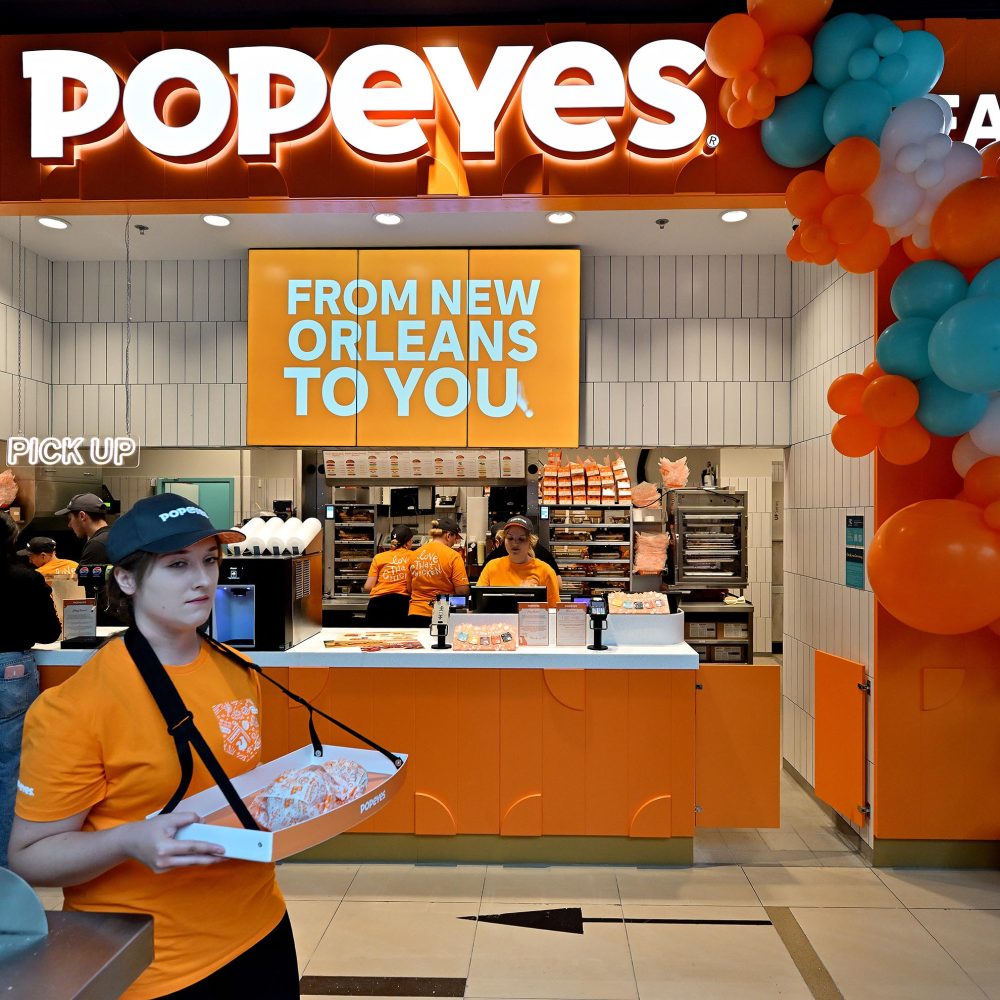 Zdjęcie przedstawia nowoczesną ladę restauracji Popeyes z dużym, podświetlanym szyldem oraz pomarańczowym ekranem z napisem „FROM NEW ORLEANS TO YOU”. Na pierwszym planie stoi pracownica w pomarańczowej koszulce i czapce z daszkiem, trzymająca na pasku tackę z zapakowanym jedzeniem, a w tle widać innych pracowników oraz dekoracje z balonów.
