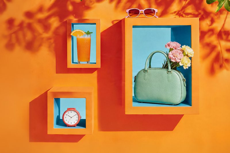 Trzy pomarańczowe ramki w formie nisz na intensywnie pomarańczowej ścianie z cieniami liści. Wewnątrz nisz znajdują się: szklanka soku pomarańczowego, czerwony budzik oraz miętowa torebka z kwiatami.