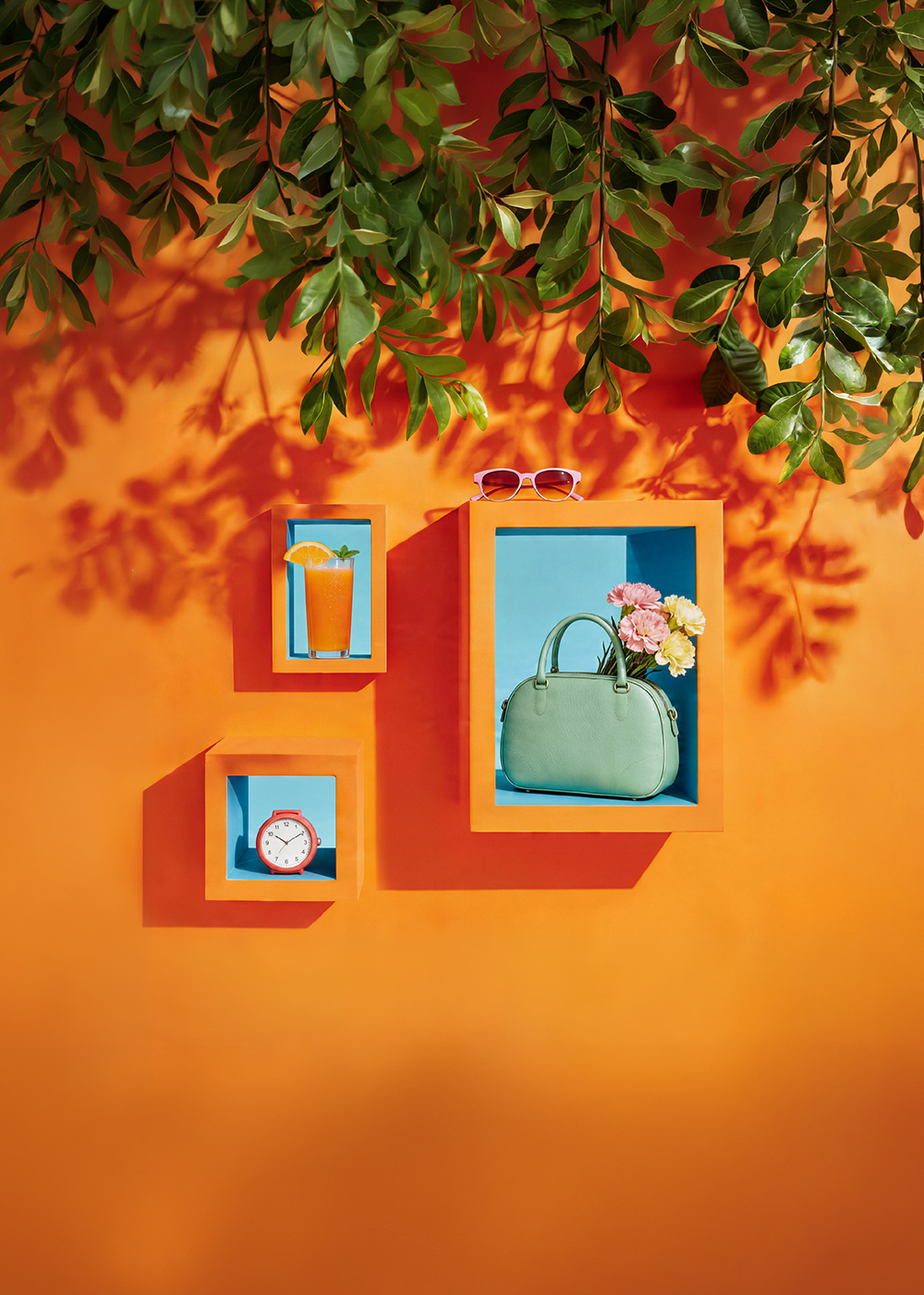Artystyczna aranżacja ścienna w kolorach pomarańczu i błękitu z letnimi akcesoriami: okularami, napojem, zegarem i torebką z bukietem.