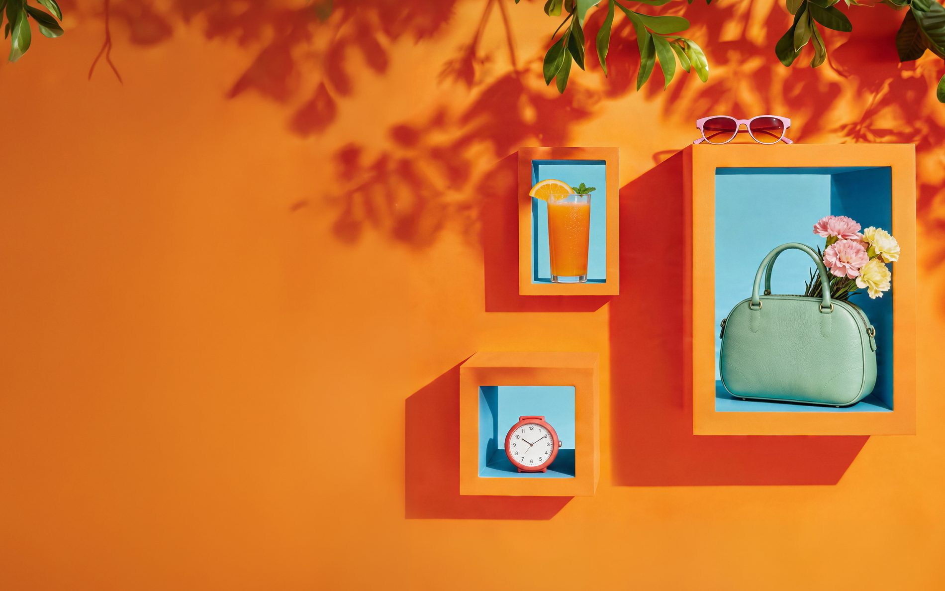Letnia kompozycja z pomarańczowymi ramami, w których wyeksponowano sok pomarańczowy, budzik i torebkę z kwiatami na tle cieni liści.