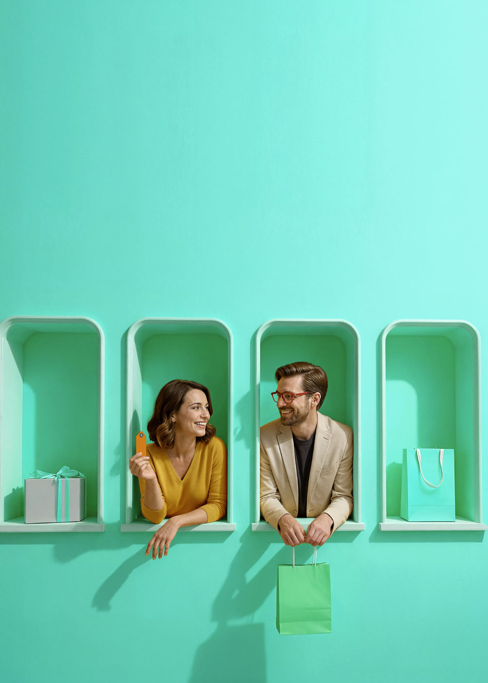 Kobieta z kartą płatniczą i mężczyzna z torbą zakupową wychylają się z dekoracyjnych turkusowych wnęk; obok ustawione są torba zakupowa oraz pudełko prezentowe.