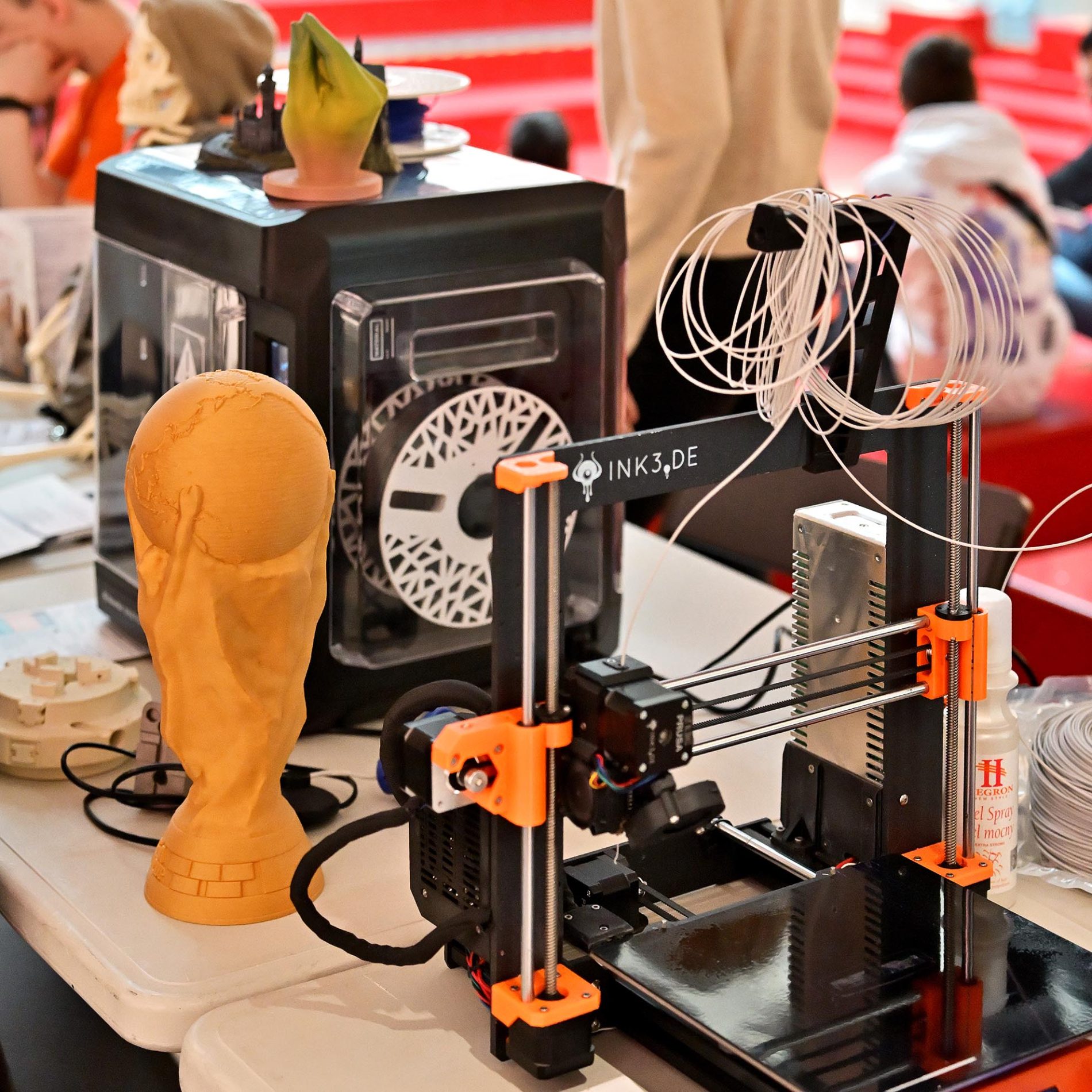 Modele 3D, w tym żółty puchar, prezentowane obok pracujących drukarek podczas wydarzenia Echo Nauki w Galerii Echo.