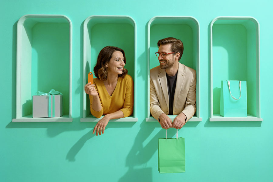 Kobieta trzymająca kartę podarunkową i mężczyzna z torbą zakupową stoją w ozdobnych wnękach na tle miętowej ściany z prezentami po bokach.