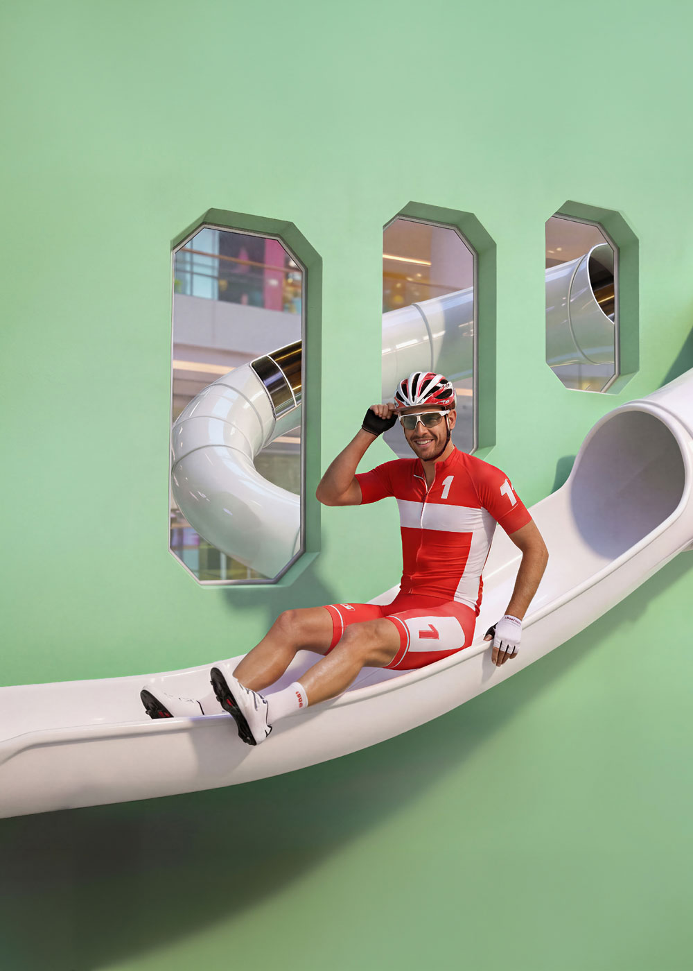 Zdjęcie przedstawia uśmiechniętego sportowca w pełnym rynsztunku kolarskim podczas nietypowej sesji na zjeżdżalni. Kontrastowa, czerwono-biała odzież kolarza odcina się od pastelowych, miętowych ścian i białych elementów konstrukcyjnych. Kompozycja jest jasna, czysta i pełna pozytywnej, zabawowej energii.