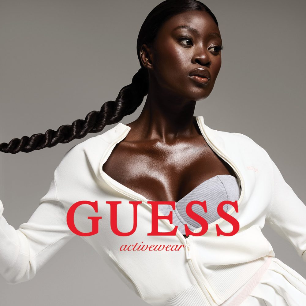 Zdjęcie promocyjne marki Guess z kolekcji Activewear, przedstawiające kobietę w fioletowym stroju sportowym podczas ćwiczeń na macie. W tle widoczny jest nowoczesny, jasny salon z dużymi oknami i minimalistycznym wystrojem. Całość uzupełnia logotyp marki Guess umieszczony w górnym rogu grafiki.