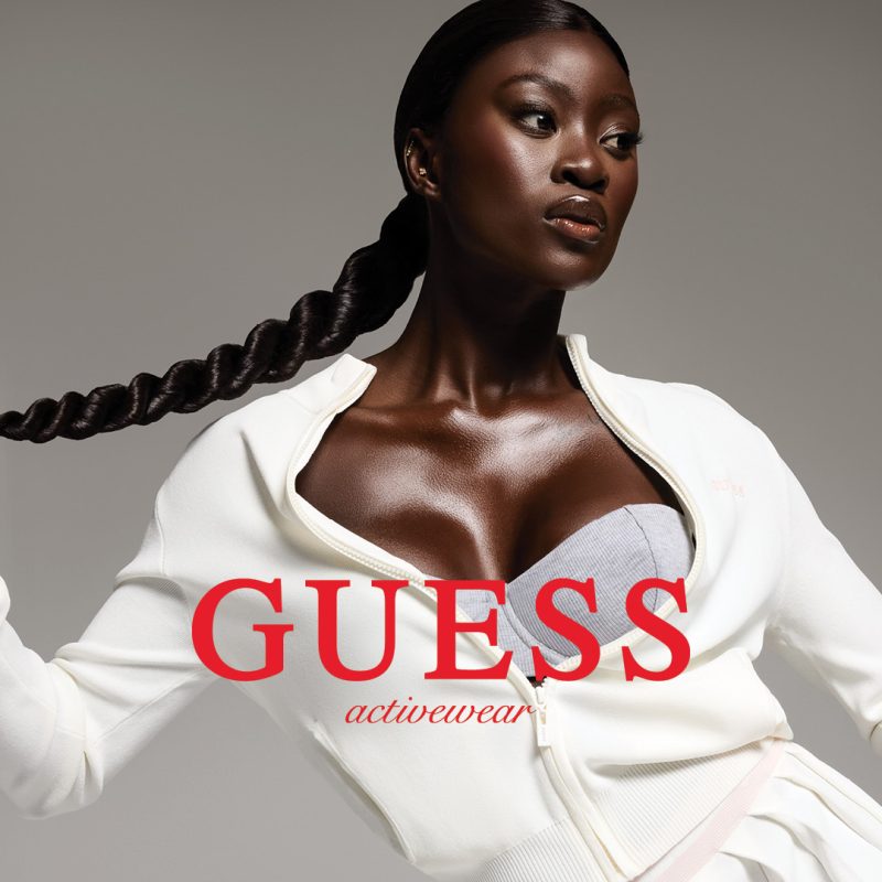 Zdjęcie promocyjne marki Guess z kolekcji Activewear, przedstawiające kobietę w fioletowym stroju sportowym podczas ćwiczeń na macie. W tle widoczny jest nowoczesny, jasny salon z dużymi oknami i minimalistycznym wystrojem. Całość uzupełnia logotyp marki Guess umieszczony w górnym rogu grafiki.
