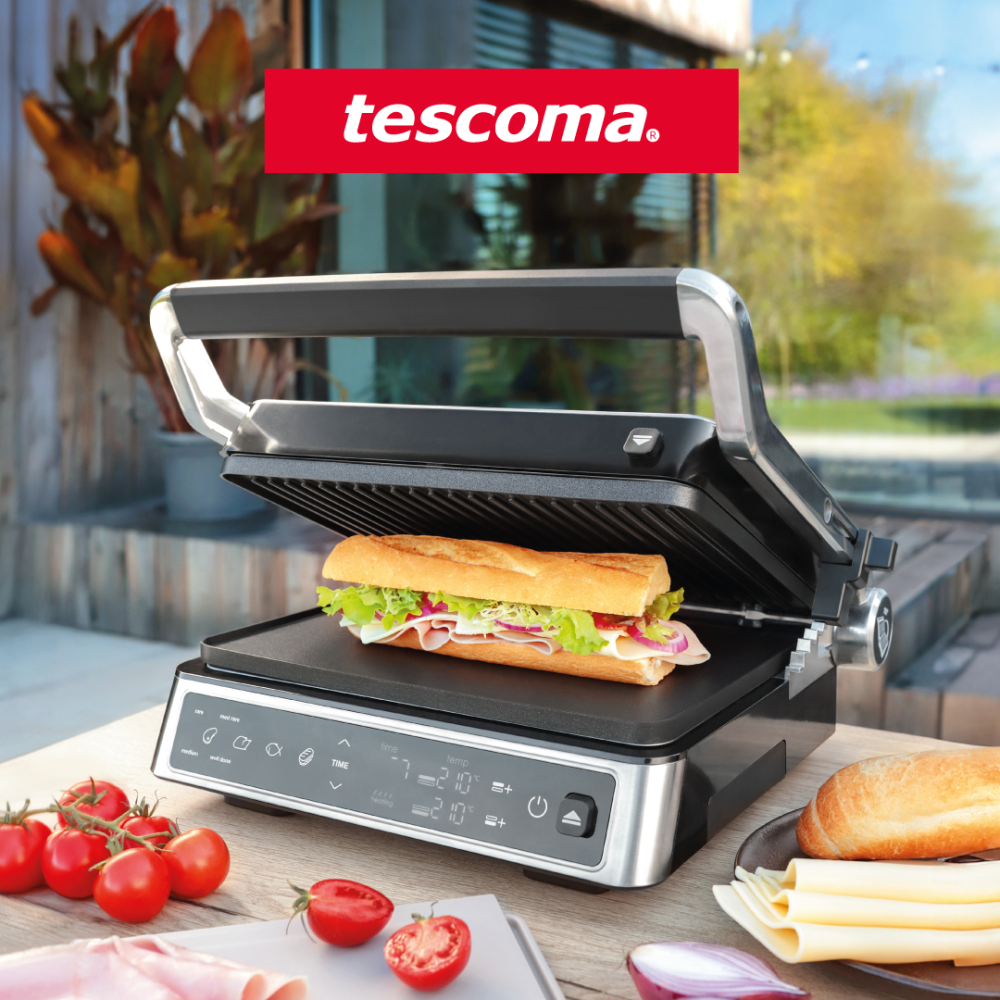 Na zdjęciu przedstawiono elektryczny grill kontaktowy marki Tescoma, na którym przygotowywana jest apetyczna kanapka z bagietki, szynki, sera oraz świeżych warzyw. Urządzenie ustawione jest na stole na tarasie, a w tle widoczny jest nowoczesny ogród, podkreślający letni, relaksujący klimat sceny. Obok grilla znajdują się świeże składniki, takie jak pomidory, cebula, ser i bagietka, tworząc estetyczną i kulinarną aranżację.
