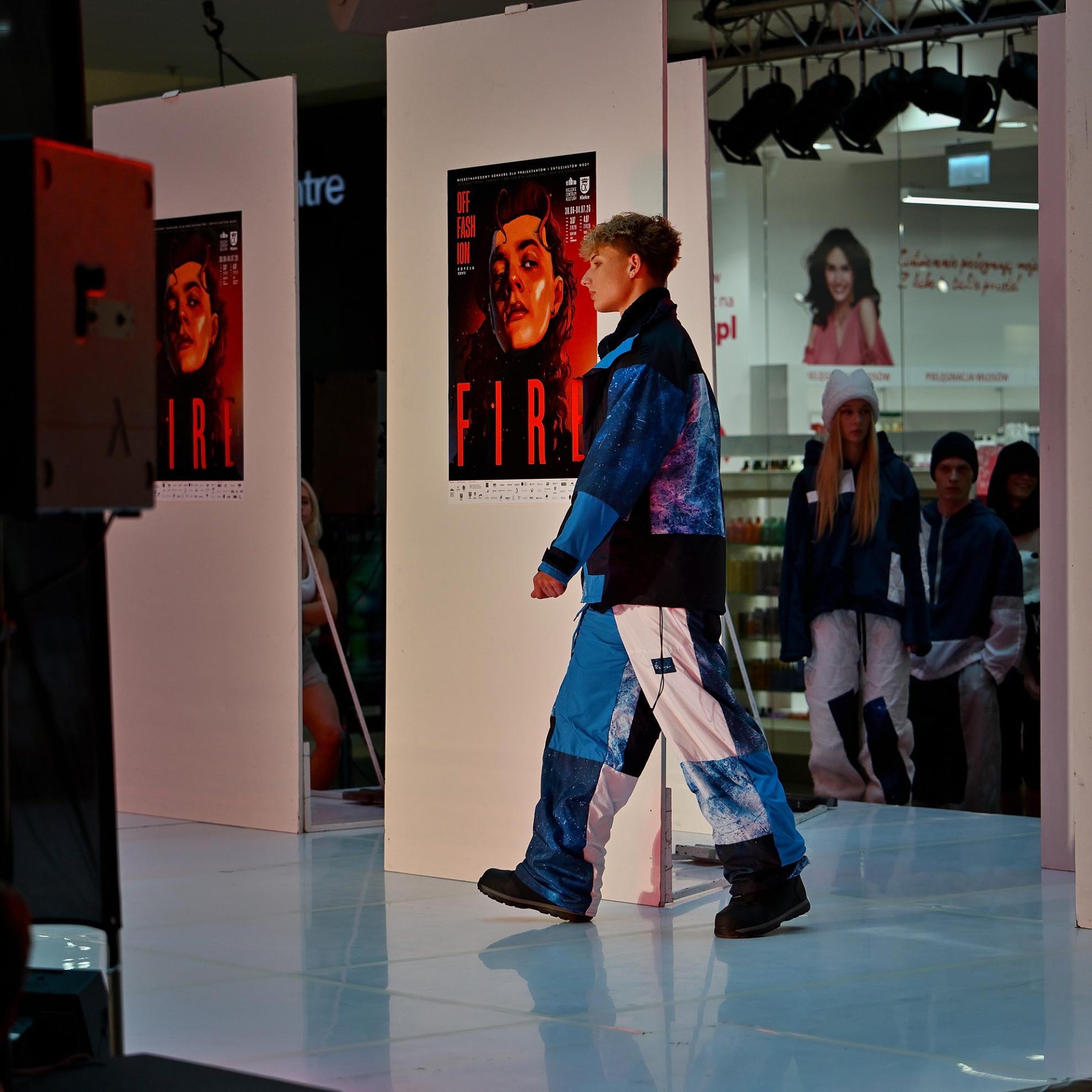 Model w niebiesko-białej stylizacji inspirowanej odzieżą techniczną przechodzi po wybiegu podczas pokazu mody w galerii handlowej. W tle znajdują się białe ekspozytory z plakatami wydarzenia oraz inni uczestnicy pokazu oczekujący na wejście na scenę. Całość oświetlona jest scenicznym światłem, a na podłodze widoczne są odbicia sylwetek i elementów scenografii.