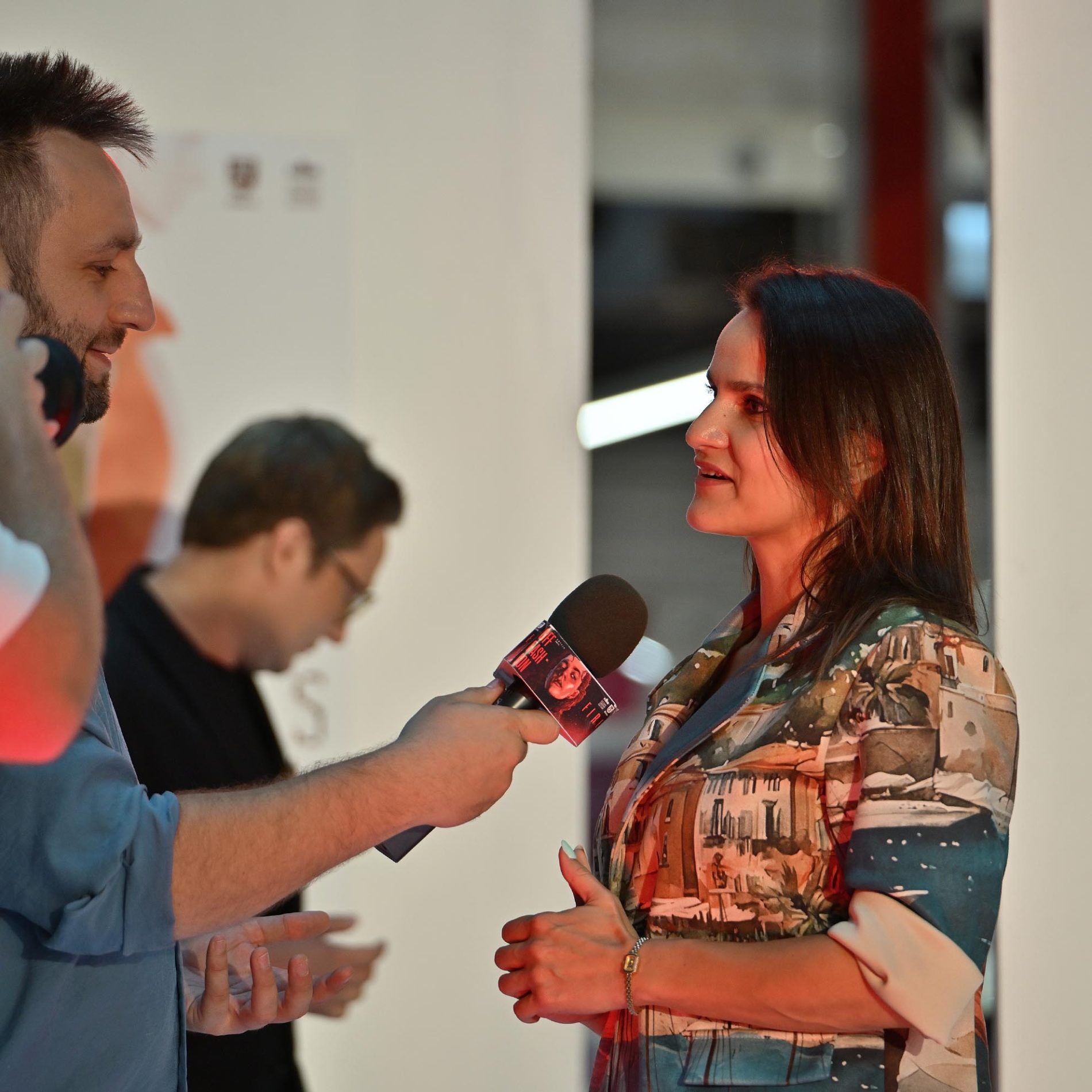 Kobieta w kolorowej marynarce rozmawia z mężczyzną trzymającym mikrofon, udzielając mu wywiadu. W tle widać zarysowaną sylwetkę innej osoby oraz fragmenty jasnego otoczenia.