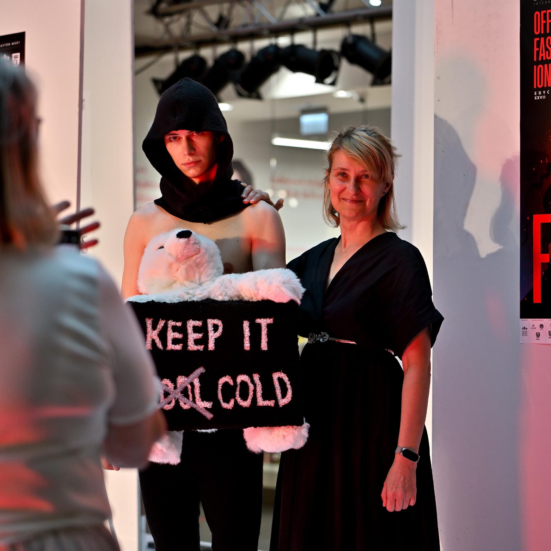 Model w czarnym kapturze trzyma maskotkę niedźwiedzia z napisem „KEEP IT COLD”, pozując obok uśmiechniętej kobiety w czarnej sukience. Scena rozgrywa się w jasnym wnętrzu, gdzie inna osoba robi im zdjęcie.