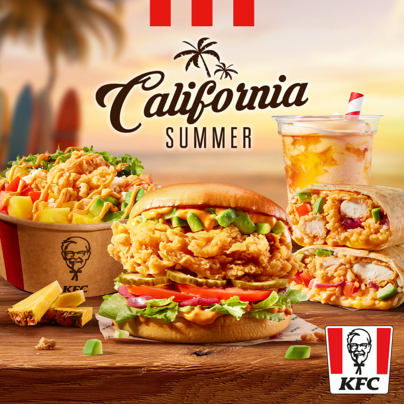 Grafika reklamowa marki KFC prezentuje letnie menu „California Summer” z burgerem, wrapem, kubełkiem pełnym chrupiącego kurczaka i owoców oraz orzeźwiającym napojem na tle plaży o zachodzie słońca. Na drewnianym stole obok dań ułożono kawałki ananasa i awokado, a nad całością widnieje stylizowany napis promocyjny oraz logo sieci.