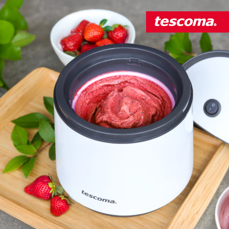 Grafika reklamowa marki Tescoma przedstawia domową maszynkę wypełnioną świeżo przygotowanym, czerwonym sorbetem truskawkowym. Urządzenie stoi na drewnianej tacy w otoczeniu świeżych truskawek oraz zielonych listków.