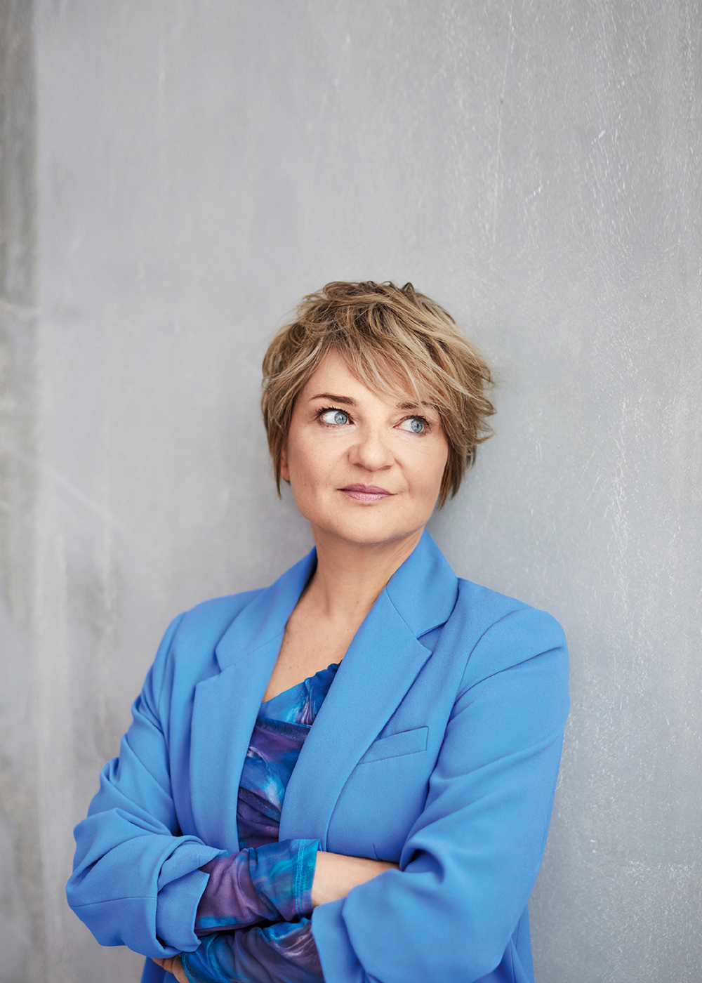 Portret kobiety w niebieskiej marynarce, stojącej z założonymi rękami na tle szarej ściany.