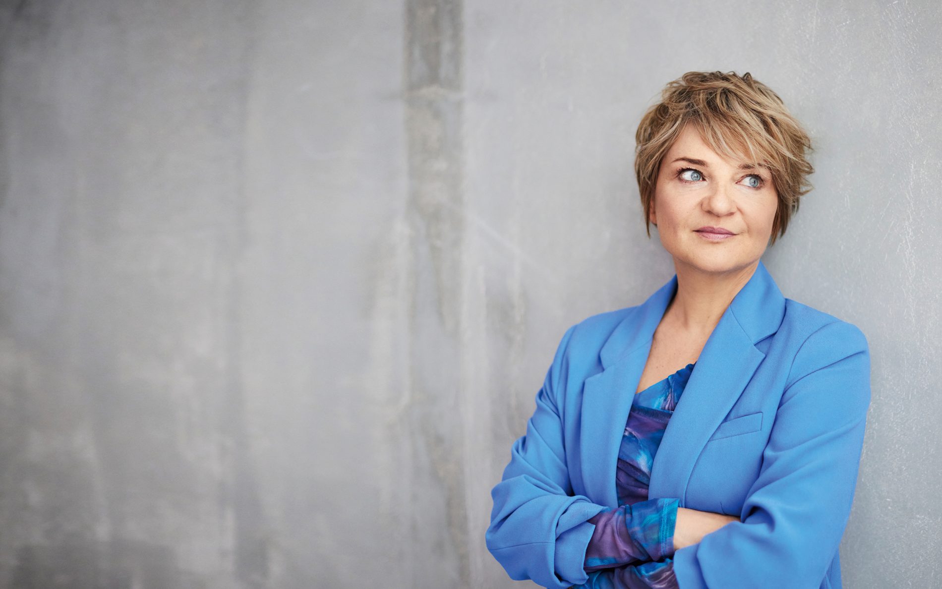 Portret kobiety w niebieskiej marynarce, stojącej z założonymi rękami na tle szarej ściany.
