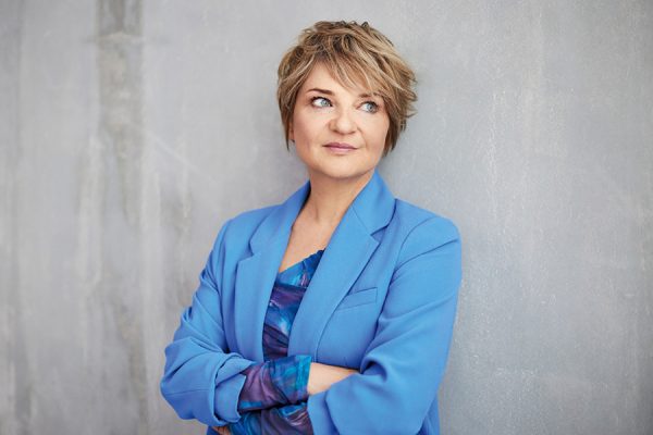 Portret kobiety w niebieskiej marynarce, stojącej z założonymi rękami na tle szarej ściany.