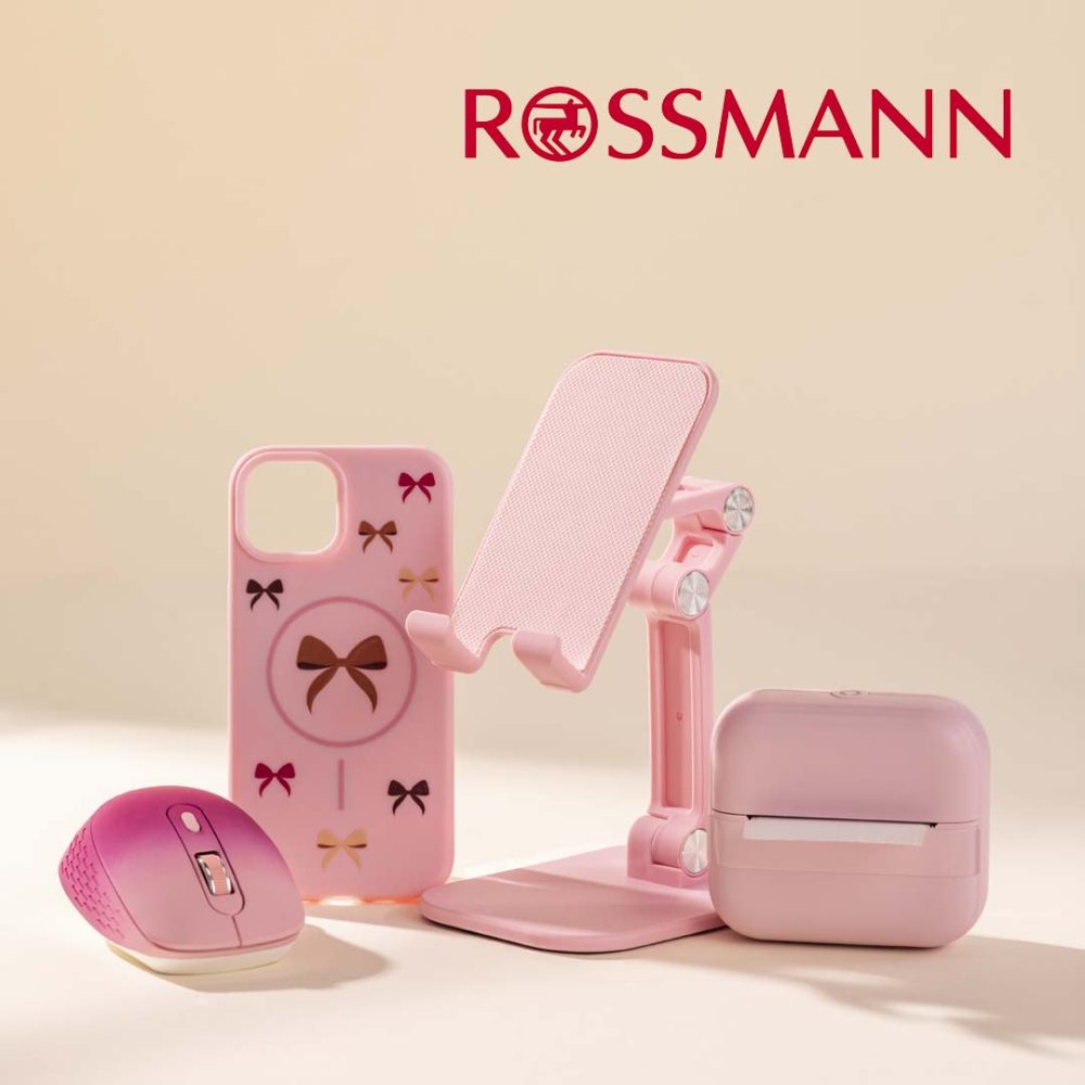 Różowe akcesoria biurkowe i elektroniczne z drogerii Rossmann: myszka komputerowa, etui na telefon w kokardki, składany stojak na telefon oraz mała drukarka termiczna. W prawym górnym rogu widnieje czerwone logo Rossmann.