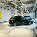 Wnętrze nowoczesnego salonu samochodowego o industrialnym charakterze, z widocznym sufitowym systemem oświetlenia i instalacji. Na jasnej podłodze wyeksponowano zielonego SUV-a marki Geely, a w głębi stoi kolejny pojazd. Po lewej stronie na białej ścianie wisi duży ekran wyświetlający abstrakcyjną, żółto-zieloną grafikę.