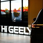 Zbliżenie na tył nowoczesnego samochodu marki Geely znajdującego się w salonie, z widocznym podświetlanym napisem „HI GEELY” na pierwszym planie. W tle przez przeszkloną witrynę widać otoczenie budynku.