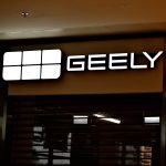 Szyld nad wejściem do salonu motoryzacyjnego marki Geely. Po lewej stronie znajduje się podświetlony na biało logotyp składający się z sześciu kwadratowych modułów ułożonych w dwa rzędy, a obok widnieje biały napis z nazwą marki „GEELY”. Poniżej widoczna jest zamknięta, ciemna roleta zabezpieczająca witrynę sklepu