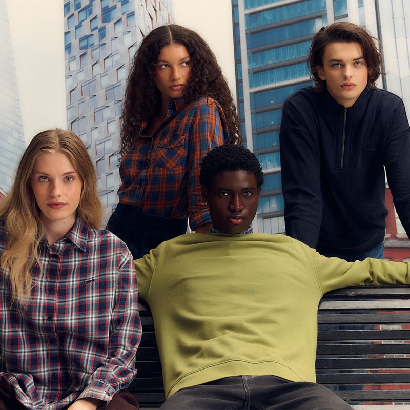 Kwadratowa fotografia wizerunkowa nowej kolekcji marki Lee Cooper. Na zdjęciu widać czwórkę młodych osób (dwie kobiety i dwóch mężczyzn) pozujących na tle wieżowca. Po lewej kobieta w koszuli w czerwoną kratę, obok kobieta w koszuli w pomarańczowo-niebieską kratę, na dole mężczyzna w oliwkowej bluzie, a po prawej mężczyzna w ciemnogranatowej bluzie zapinanej pod szyją.