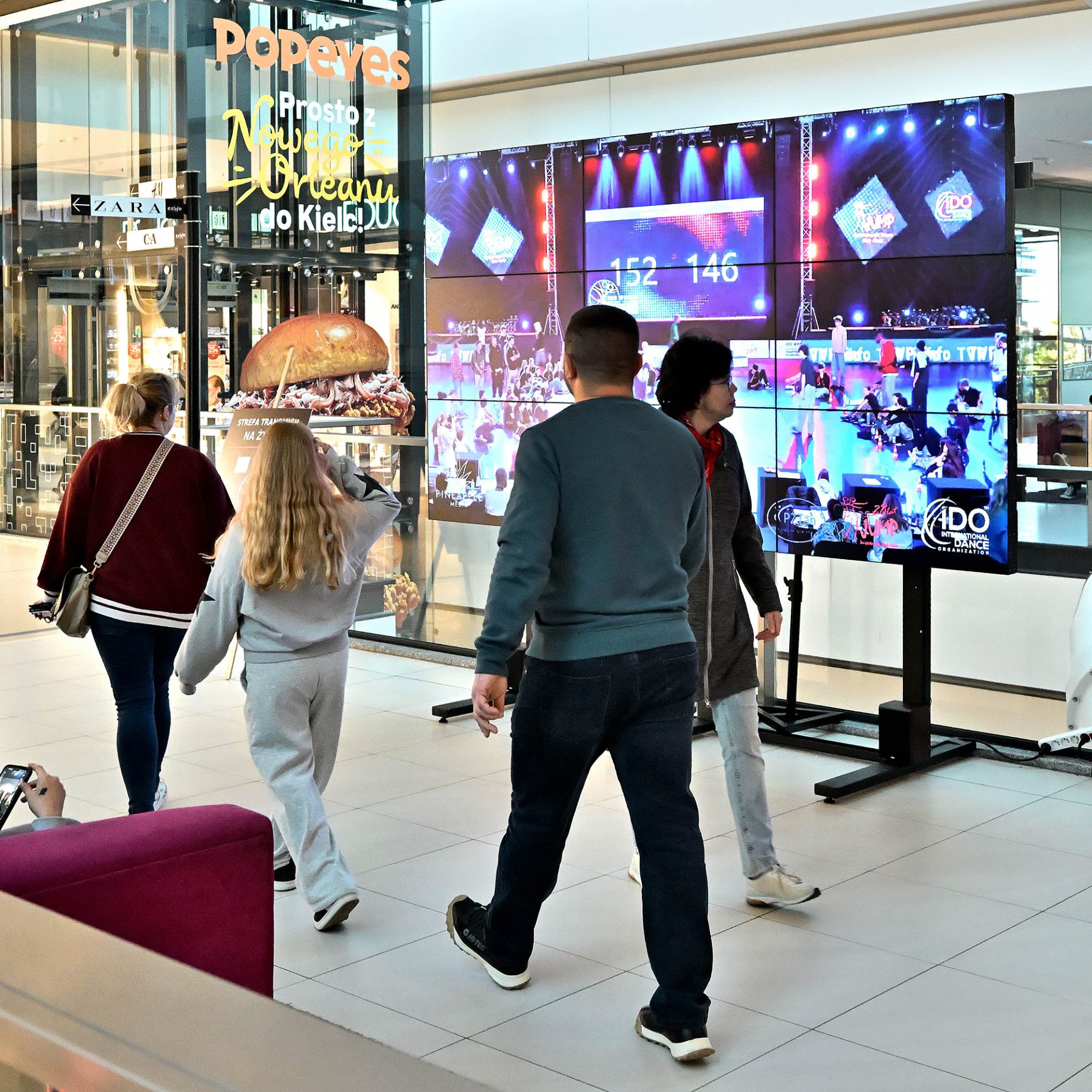 Osoby spacerujące w pasażu Galerii Echo mija wielki ekran z transmisją pokazów tanecznych.