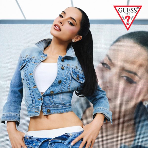 Grafika promocyjna marki Guess prezentująca kolekcję jeansową z udziałem piosenkarki Becky G. W prawym górnym rogu znajduje się charakterystyczne czerwono-białe logo marki z trójkątnym napisem „GUESS”. Na pierwszym planie pozuje modelka z ciemnymi włosami spętnymi w wysoki kucyk, ubrana w krótki biały top, dżinsową kurtkę ze zdobieniami z dżetów oraz dżinsy z obniżonym stanem. W tle widoczny jest wielkoformatowy ekran z powiększonym portretem artystki.